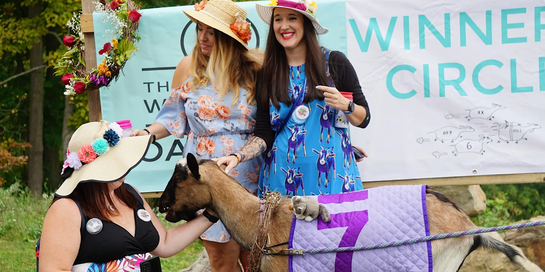 Three women in sun hats stand with a donkey wearing a purple quilted blanket at an outdoor event with a banner in the background.