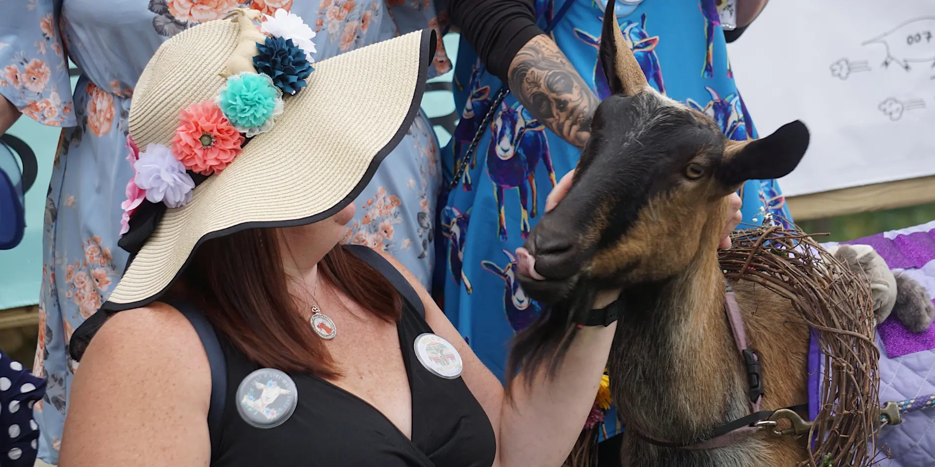 Woman in a flower-adorned sun hat pets a brown donkey at a festival, with people in bright outfits nearby.