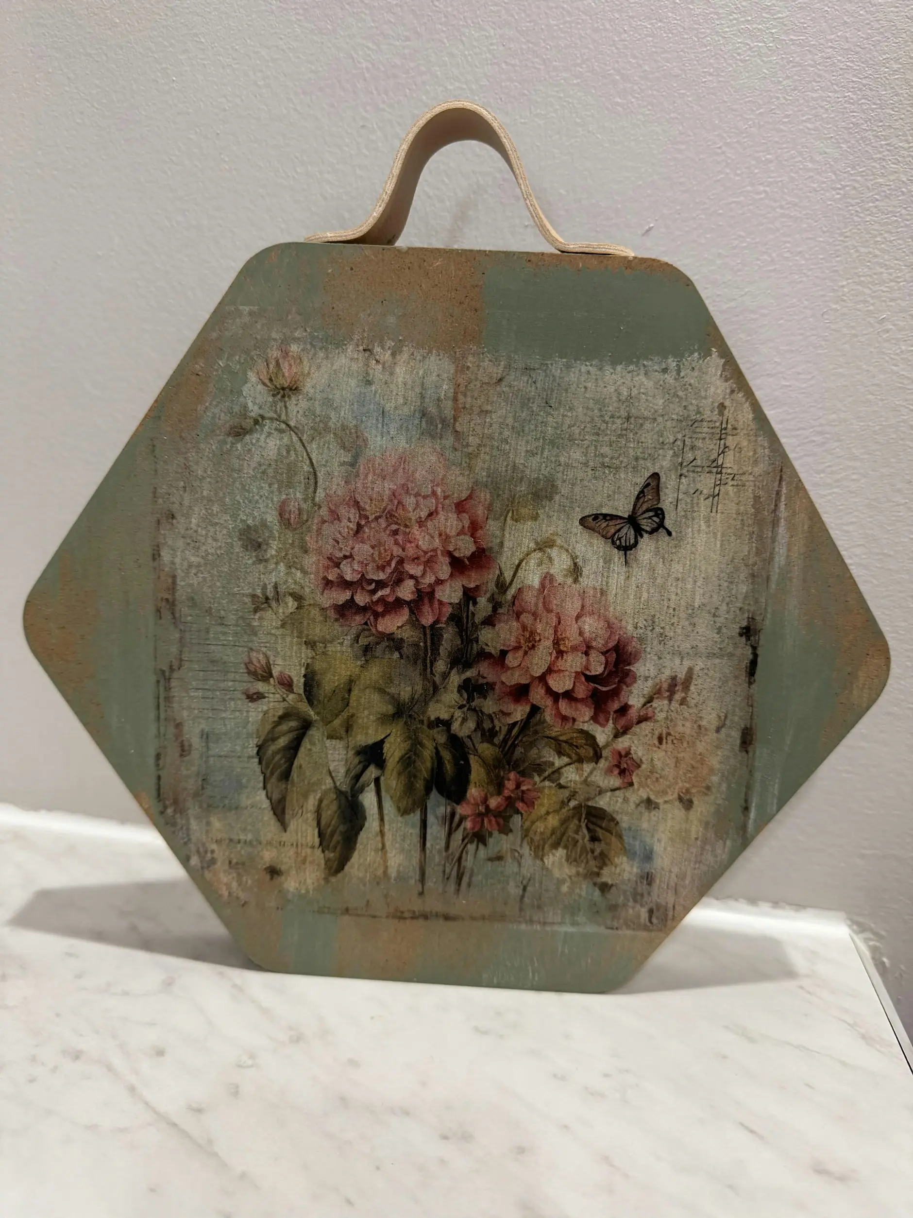 Decorative, vintage-style plaque with pink flowers and a butterfly on a weathered background, hanging by a leather handle on a wall.