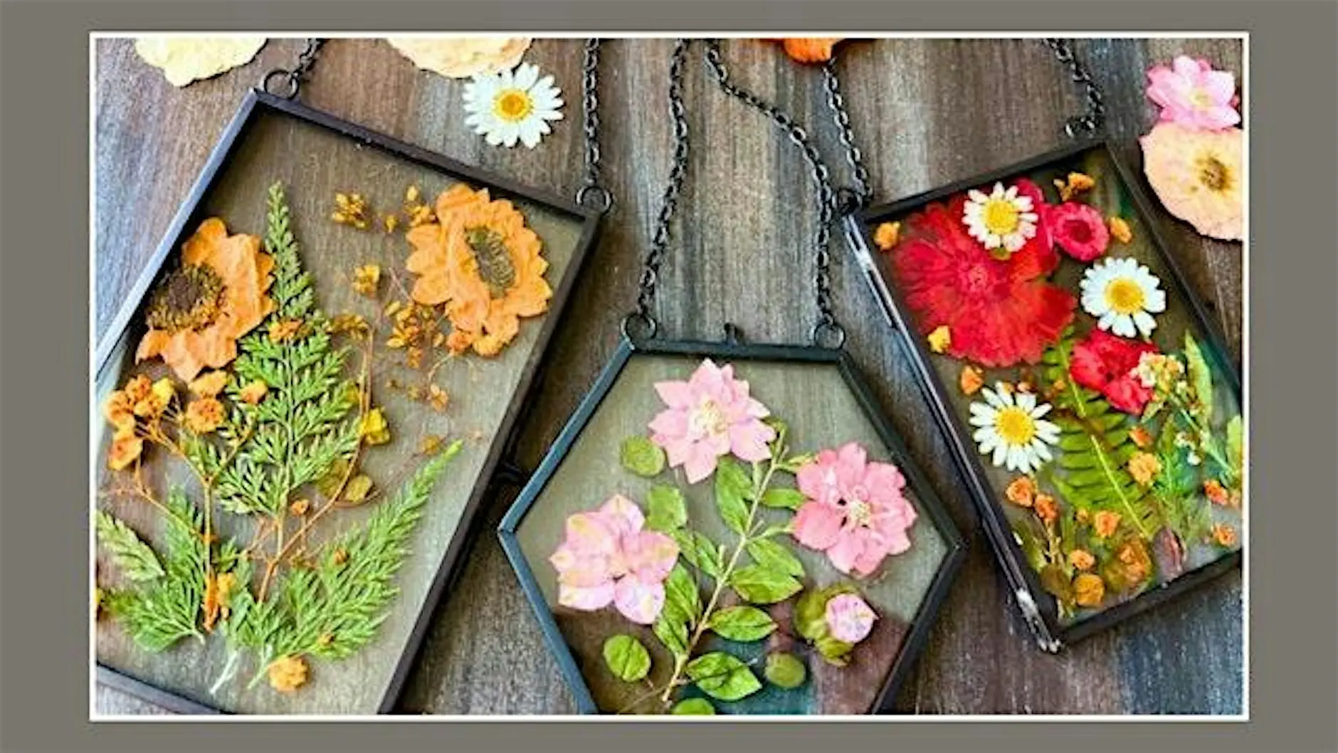 Geometric glass frames with pressed flowers in orange, pink, and white on a weathered wooden surface. The frames are linked by dark metal chains.