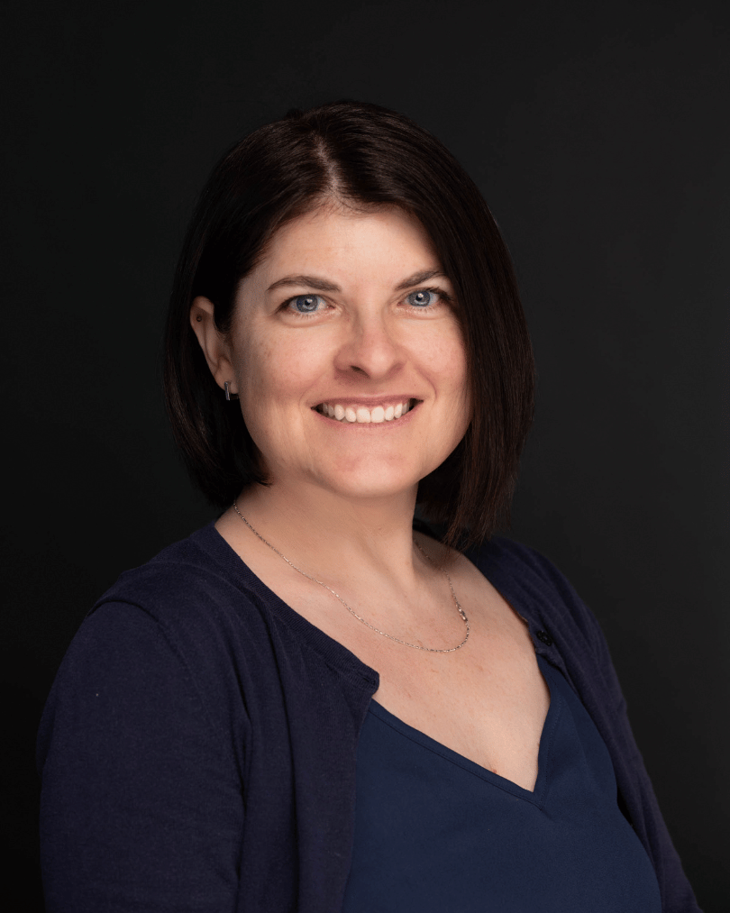 Portrait of a smiling woman with dark hair in a navy blouse and cardigan against a dark background, head-and-shoulders shot.